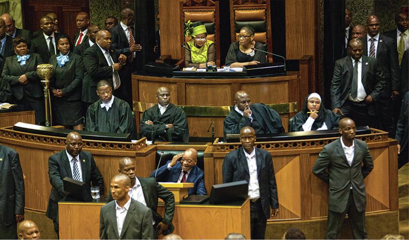 The former president of South Africa laughing while chaos envelops Parliament and angry voices fill its chambers.
